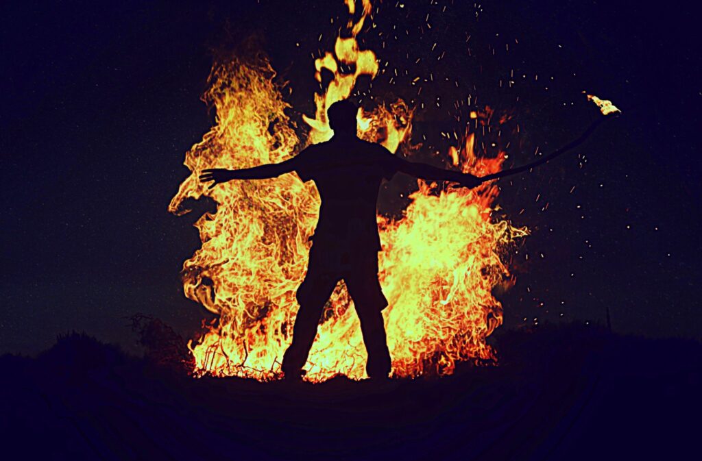 Mann im Dunkeln als Silhouette vor einem Lagerfeuer mit Fackel in der Hand.